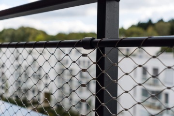 Černé zábradlí s drátěnou sítí na balkoně YIT Parvi Cibulka, v pozadí rozmazané bílé budovy a stromy.