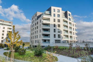 Moderní obytná budova v Britské Čtvrti s prosklenými balkony obklopená zelení pod modrou oblohou.