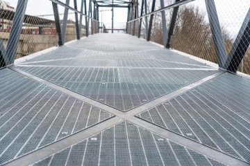 Ocelová pěší lávka s kovovým zábradlím a výhledem na železniční trať.