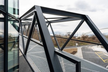 Ocelová lávka s moderním designem propojující dvě budovy Port 7, s prosklenou konstrukcí a výhledem na silnici.