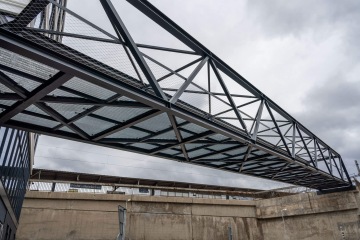 Ocelová pěší lávka nad betonovým nádražím proti zamračené obloze.