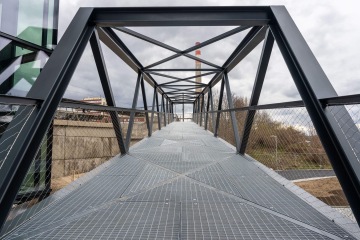 Moderní kovová lávka Port 7 s mřížovanou podlahou a geometrickou konstrukcí pod zamračenou oblohou.