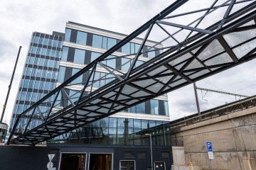 Skleněná budova Port 7 propojená moderní lávkou nad silnicí s matnou oblohou v pozadí.