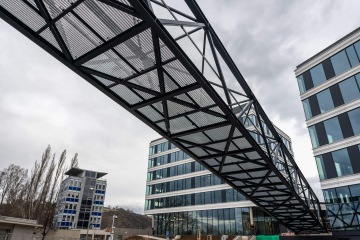 Kovový most spojuje dvě moderní skleněné budovy Port 7 na pozadí zamračené oblohy.