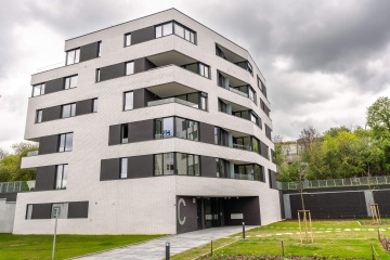 Moderní pětipatrová budova Rezidence U Sluncové s bílými cihlami a tmavými okny vedle zeleného trávníku pod zamračenou oblohou.