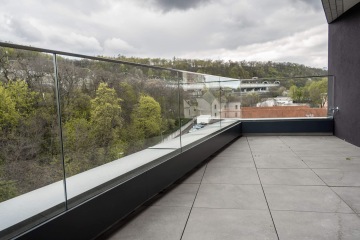 Terasa Rezidence U Sluncové s proskleným zábradlím s výhledem na stromy a budovy pod zamračenou oblohou.