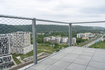 Pohled z terasy Vanguard Praha přes drátěné zábradlí na moderní bytové domy obklopené zelení a silnicí s lesnatými kopci v pozadí.