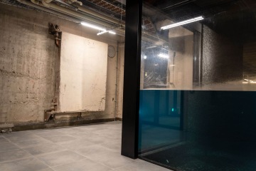 Podzemní místnost s betonovými stěnami, prosklenou stěnou a nasvětleným potrubím ve Vanguard Praha.