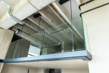 Skleněný balkón zavěšený na kovových tyčích v moderním interiéru Vanguard Praha s viditelnými potrubími na stropě.