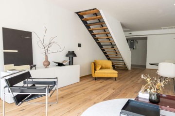 Moderní interiér Rezidence Erbenova s žlutým křeslem, černou kovovou židlí, dřevěnými schody a minimalistickou dekorací na bílém nábytku.