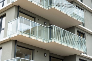 Moderní balkon v Britské Čtvrti s průhledným zábradlím a vzorovaným panelem, šedá fasáda budovy.