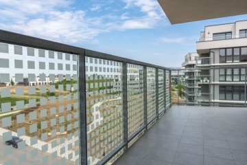 Prostorný balkon s moderním skleněným zábradlím, výhled na nové bytové domy v Britské Čtvrti a modrou oblohu s bílými mraky.