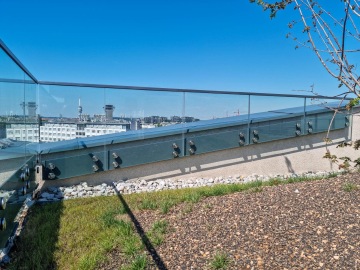 Celoskleněné zábradlí na střešní terase s výhledem na město pod modrou oblohou, okolní zeleň a kameny.