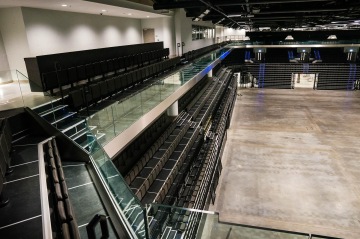 Prázdná koncertní hala s černými sedadly, celoskleněným zábradlím a betonovou podlahou.