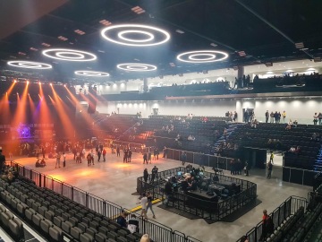 Osvětlený koncertní sál s kruhovými světly na stropě, celoskleněným zábradlím a lidmi shromažďujícími se před pódiem.