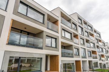 Moderní bytový dům s balkony s celoskleněným zábradlím a šedobílou fasádou pod zataženou oblohou.
