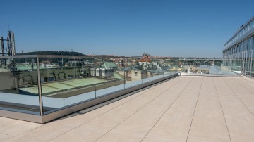 Střešní terasa s celoskleněným zábradlím a výhledem na centrum Prahy pod modrou oblohou.