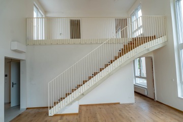 Moderní interiér Modřanský Cukrovar s bílým schodištěm a dřevěnou podlahou, velká okna zajišťují světlé prostředí.