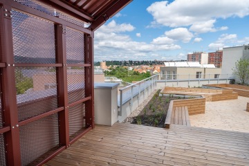 Střešní terasa Modřanský Cukrovar s dřevěnou podlahou a záhony nabízí výhled na město za slunečného dne s modrou oblohou.