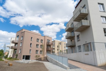 Moderní obytné budovy Modřanský Cukrovar s balkóny za slunečného dne, obklopené upraveným chodníkem a stromy.