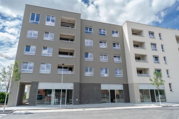 Moderní pětipatrová bytová budova Modřanský Cukrovar se skleněnými výlohami a zaparkovanými automobily, obklopená stromy pod oblohou s mraky.