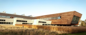 Moderní budova UCEEB s dřevěným obkladem a šikmými stěnami, obklopená suchou trávou pod modrou oblohou.