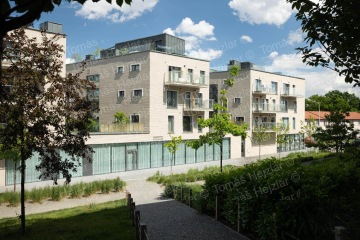 Moderní bytový dům s prosklenými balkony a zelení v popředí pod modrou oblohou.