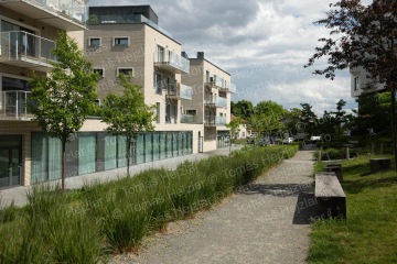 Moderní bytový dům s balkony vedle pěší cesty lemované stromy a keři.