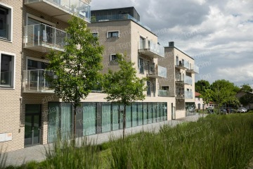 Moderní bytový dům s prosklenými balkony a zelení před ním za polojasného dne.
