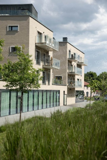 Moderní bytový dům s balkony a proskleným přízemím, obklopený zelení a stromy pod zamračenou oblohou.