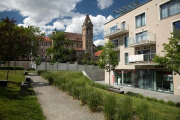 Moderní budova s balkóny vedle historické věže, stromy a pěšinou v parku za slunečného dne.