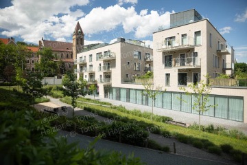 Moderní bytový dům s balkony vedle historického kostela, obklopený udržovanou zelení.