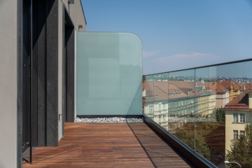Moderní terasa s dřevěnou podlahou, skleněným zábradlím a dělící stěnou, s výhledem na budovy s červenými střechami pod modrou oblohou.