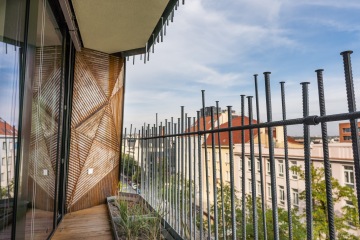 Balkon s kovovým zábradlím a atypickou dělící stěnou, s výhledem na město za slunečného počasí.
