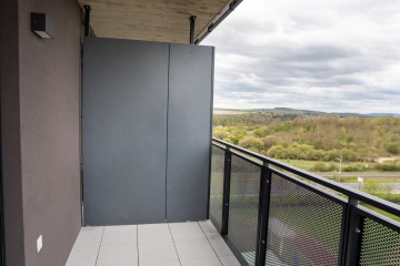 Balkon s kovovým zábradlím, dělící stěnou a pohledem na zelenou krajinu pod zamračenou oblohou.