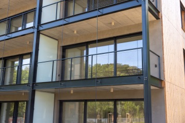 Moderní budova s prosklenými balkony, dělícími stěnami a černým kovovým zábradlím, odrážející okolní zeleň.