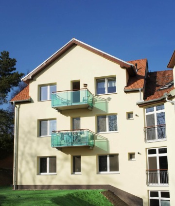 Žlutý bytový dům Dolní Kounice se skleněnými balkony a červenou střechou za slunečného dne.