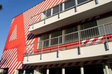Budova s červeno-bílými diagonálními pruhy a balkony s kovovým venkovním zábradlím pod modrou oblohou.