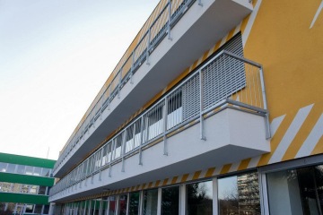 Žlutá budova s balkonem s kovovým venkovním zábradlím a pruhy na fasádě.