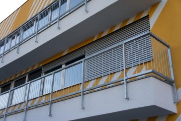 Balkon s kovovým venkovním zábradlím na moderní budově v barvách šedé a žluté.
