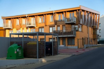 Moderní třípodlažní bytový dům s balkóny, vedle stojí recyklační kontejnery a popelnice.