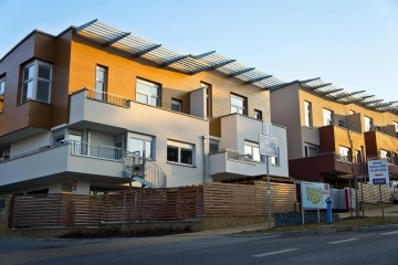 Moderní řadové domy s dřevěnými panely a balkony, dopravní značka a prodejní tabule před nimi.