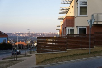 Moderní budova s dřevěným plotem, v pozadí panorama města za jasného dne.