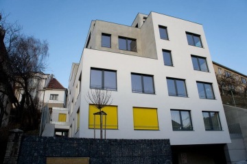 Moderní bílá budova Nad Vinohradem s velkými okny a žlutými okenicemi.