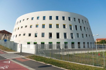 Moderní kruhová budova Centrální depozitář UPM s mnoha okny, oplocení a cyklostezka v popředí.