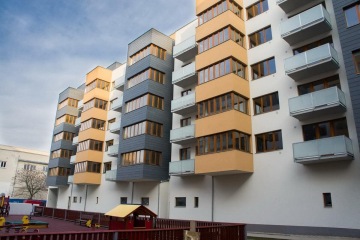 Moderní bytový dům s balkony a dětským hřištěm v popředí.