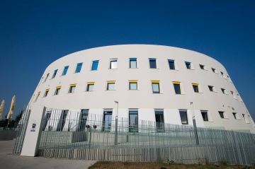 Moderní kruhová budova Centrální depozitář UPM s žlutými a modrými okenicemi za plotem pod jasnou modrou oblohou.