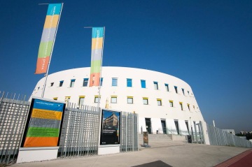 Moderní bílá budova Centrální depozitář UPM s kruhovým tvarem, barevnými banery a oplocení za slunečného dne.