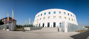 Moderní bílá kruhová budova Centrální depozitář UPM s vícebarevnými okny, vedle ní visí barevné vlajky na modré obloze.