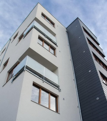 Moderní bytový dům s bílou fasádou a skleněnými balkony pod modrou oblohou.